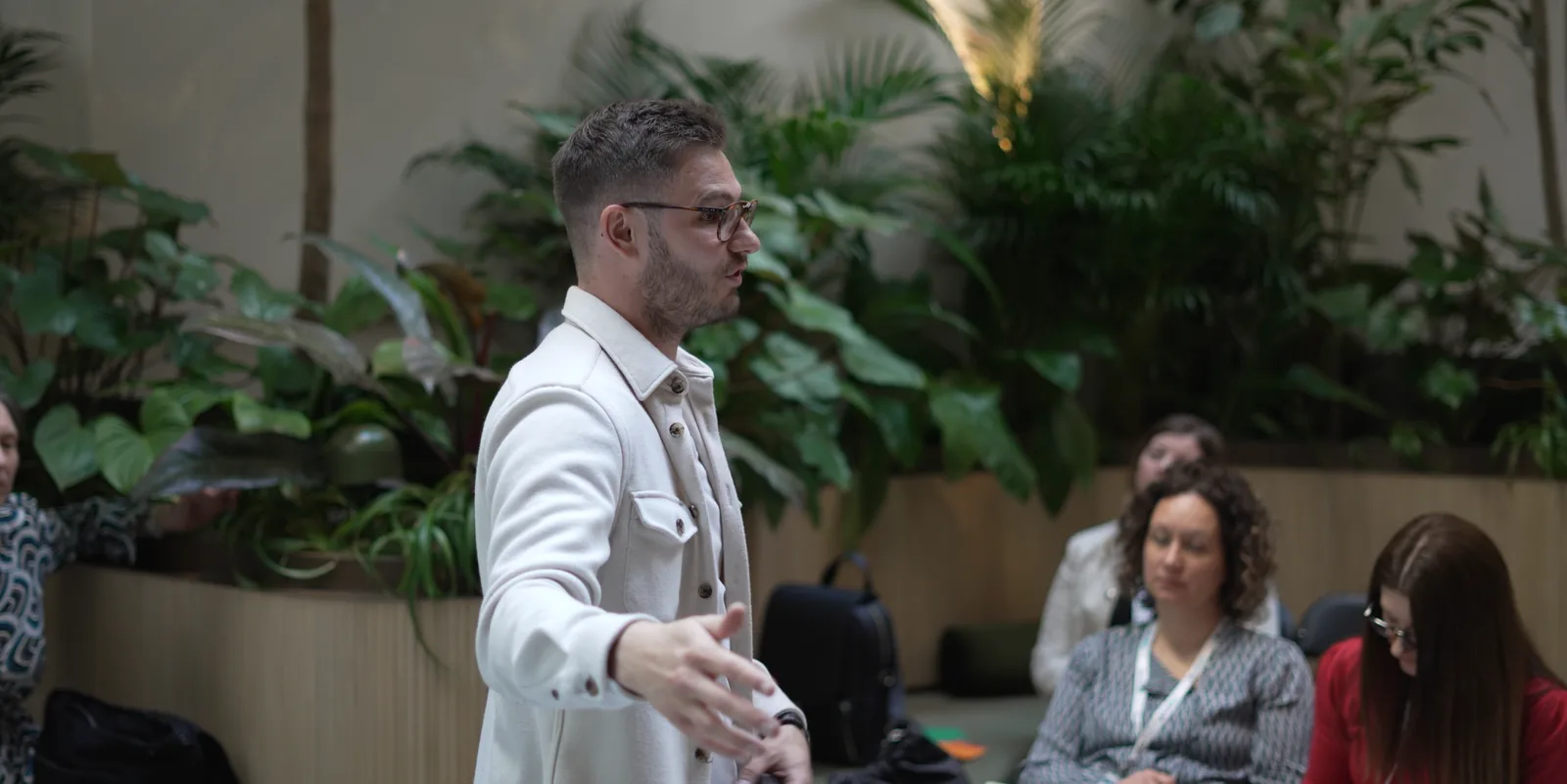 Speaker in a cream jacket addresses a seated audience against a wall of tropical plants.