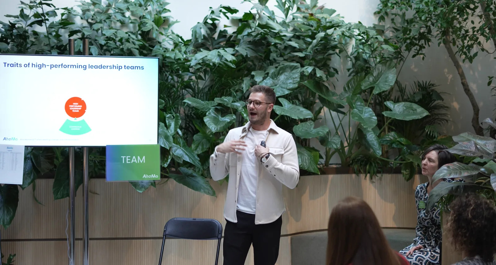 Presenter beside a screen reading 'Traits of high-performing leadership teams', plants behind.