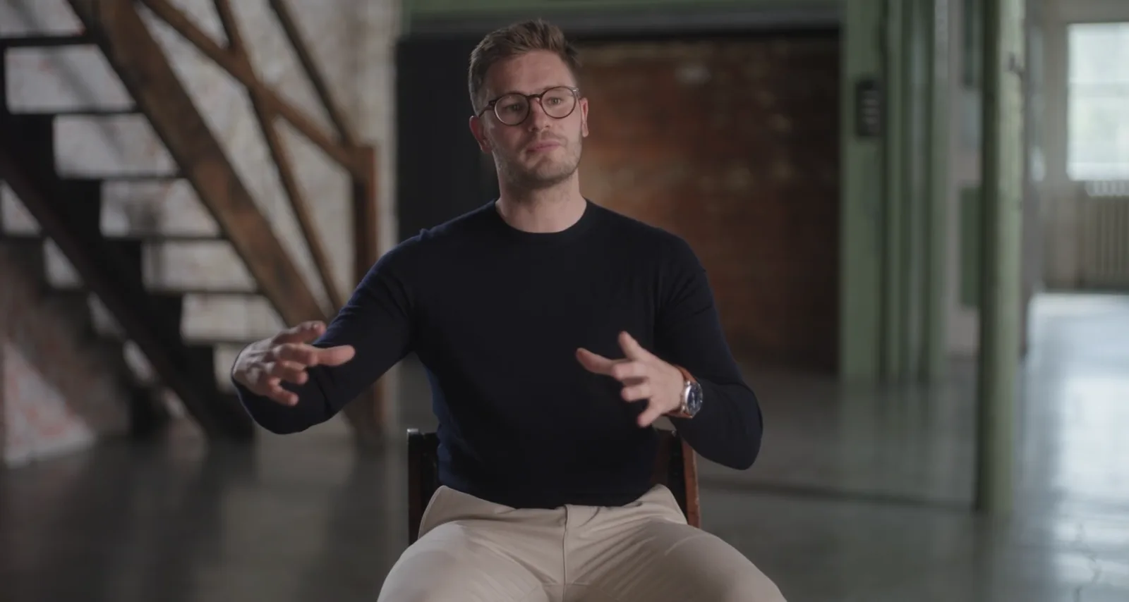 Man in a navy jumper gestures mid-sentence, seated in an exposed-brick interior with a staircase.
