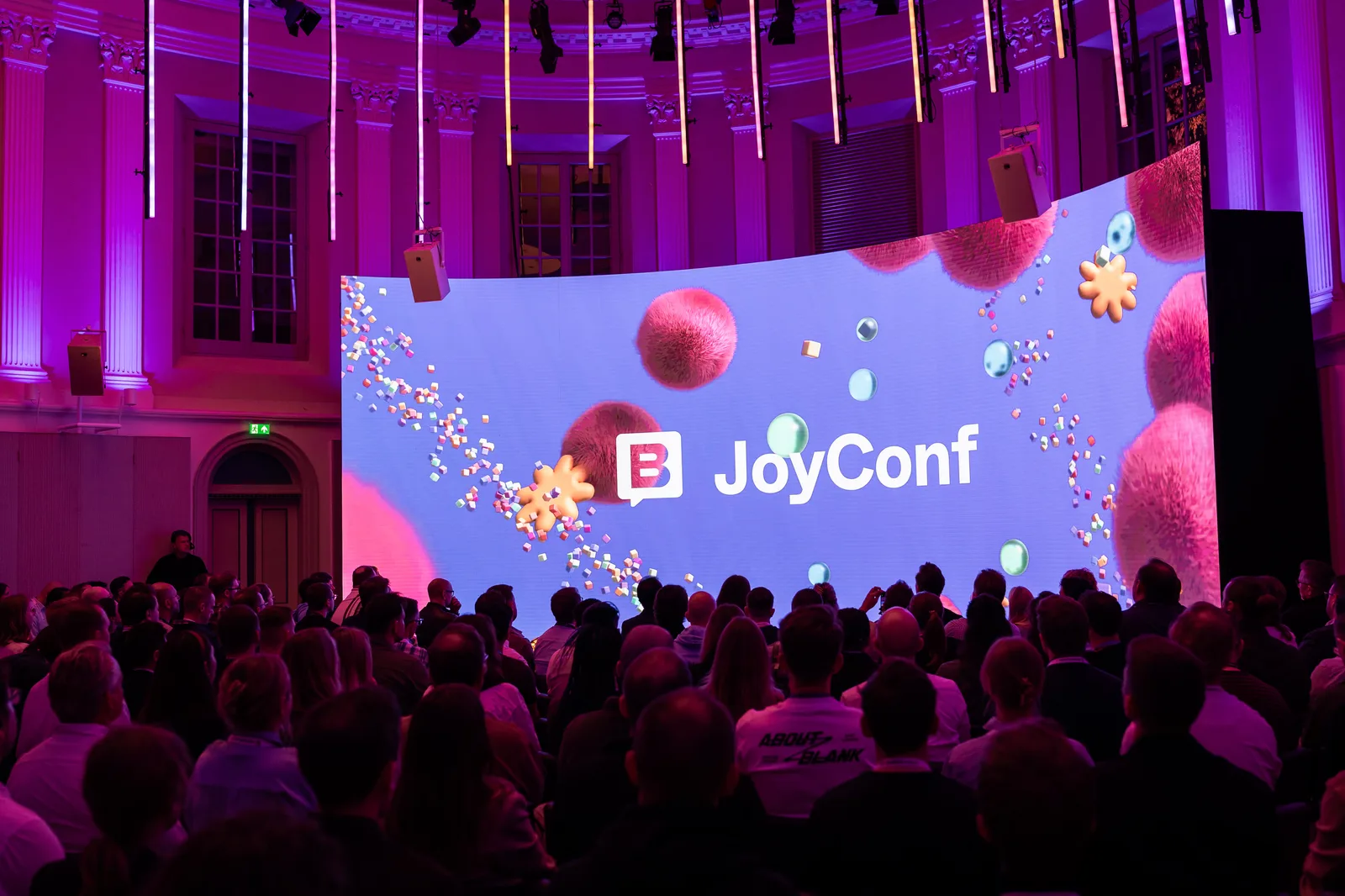 A curved LED stage screen glows purple with the JoyConf logo and giant fuzzy pink spheres above a packed seated crowd.