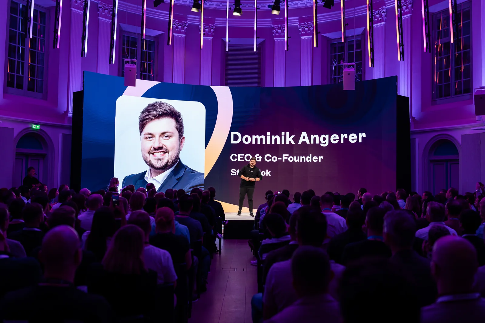 A speaker in black stands before a huge screen introducing "Dominik Angerer, CEO & Co-Founder" to a full auditorium.