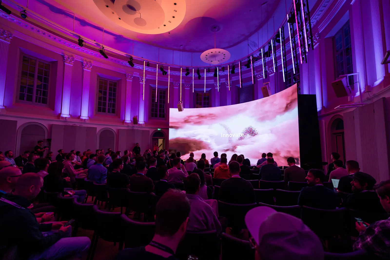 Wide view of a domed hall, the stage screen glowing with pink clouds and the word "Innovate" above rows of delegates.