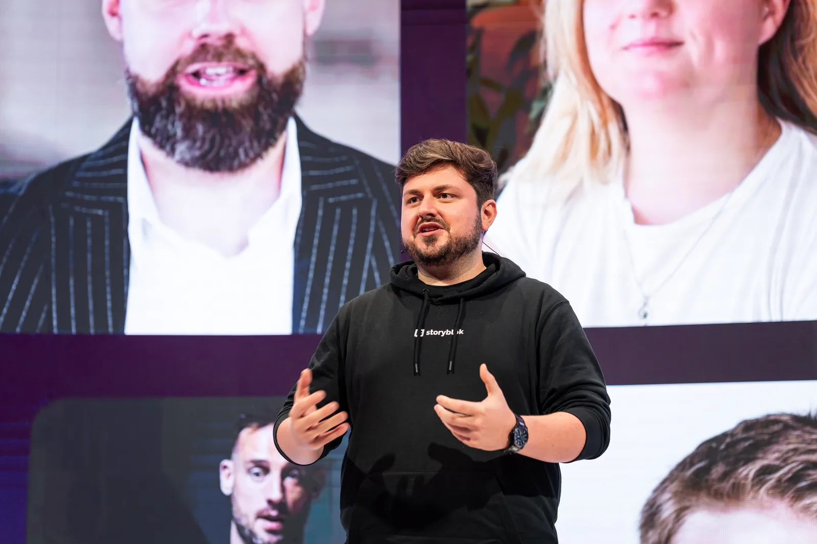 A speaker in a black Storyblok hoodie gestures mid-sentence, oversized attendee portraits lighting the screen behind him.