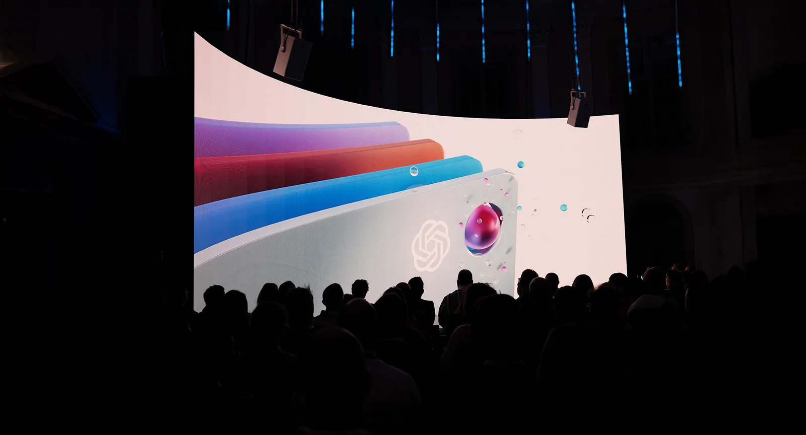 A dim auditorium, silhouetted heads watching a screen filled with layered coloured ribbons and a swirling white mark.