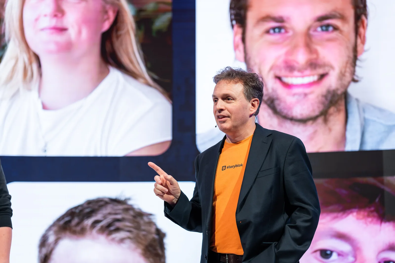 A speaker in an orange Storyblok t-shirt points mid-sentence, giant attendee portraits lighting the screen behind him.