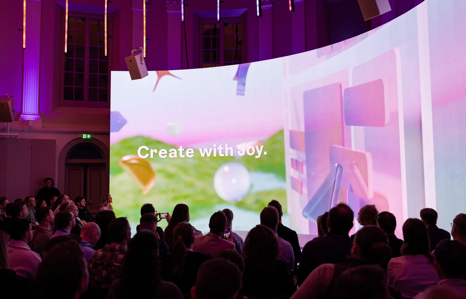 Stage screen reading "Create with Joy." over pastel 3D shapes, watched from a domed, pillared hall.
