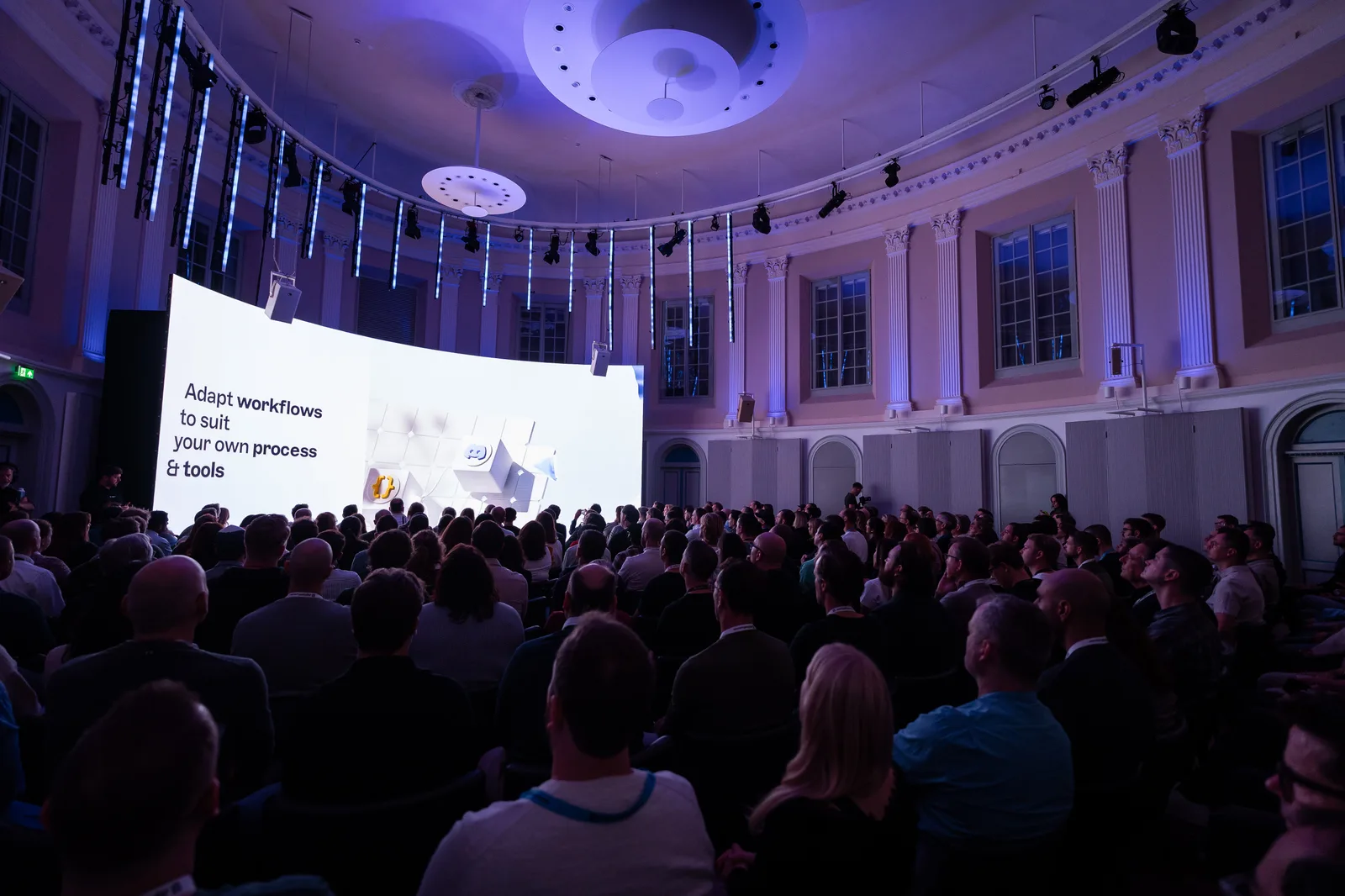 Screen text reading "Adapt workflows to suit your own process and tools" above a white 3D icon, watched by a packed hall.