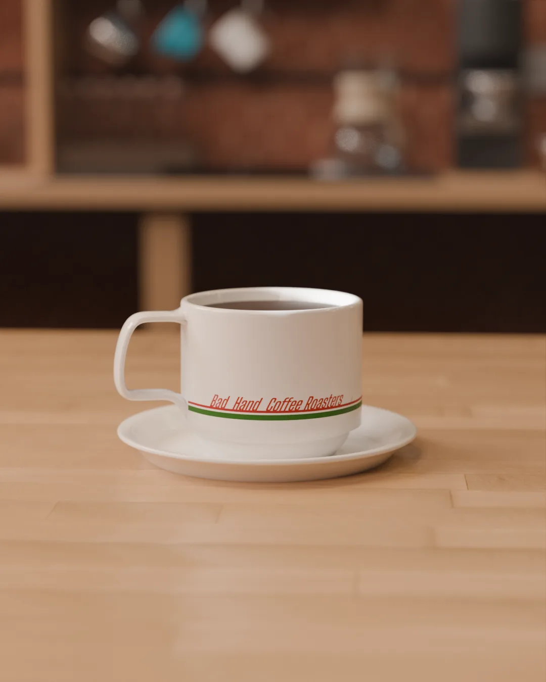 White cup and saucer banded in red and green 'Bad Hand Coffee Roasters' lettering, on a wooden counter.