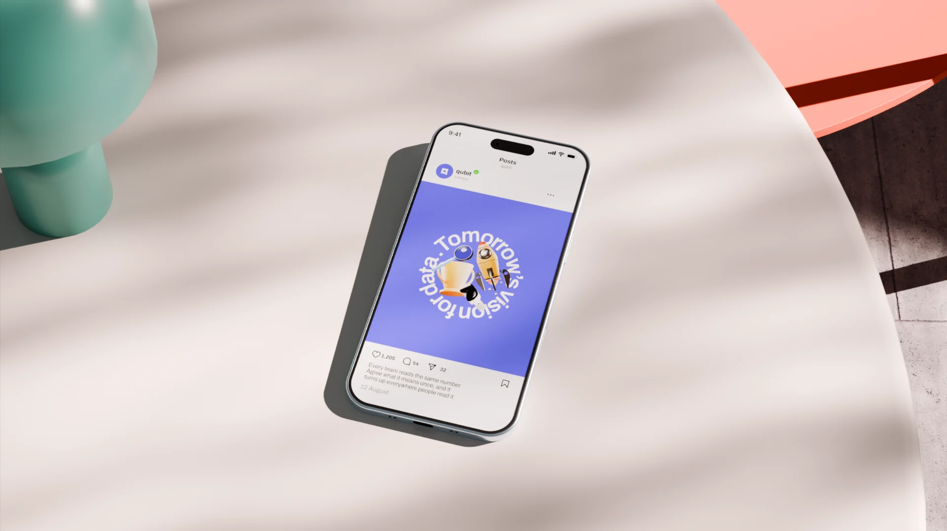 A phone lying on a pale draped surface shows a Qubit social post: a trophy and a rocket inside curved lettering on violet, with likes, comments and a caption below it.
